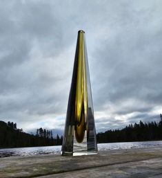 a tall metal sculpture sitting on top of a wooden platform next to a lake with trees in the background