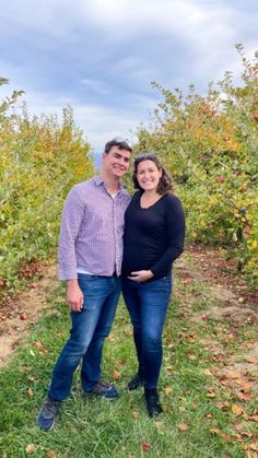 a man and woman standing next to each other in an apple tree filled field with lots of fruit