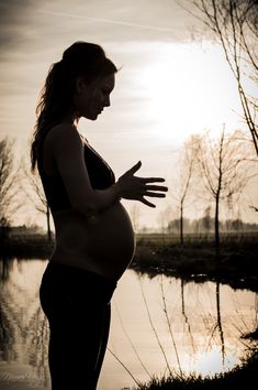 a pregnant woman standing in front of a body of water with her hands out to the side