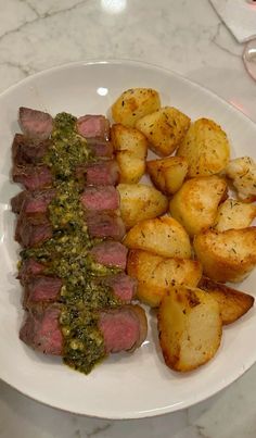 a white plate topped with meat, potatoes and pesto next to a glass of wine