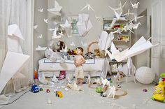 a child standing in front of a bed surrounded by toys and paper cranes hanging from the ceiling