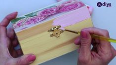 two hands holding a paintbrush and painting a wooden box with flowers on it,