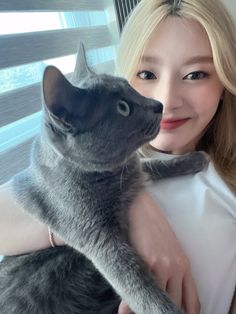 a woman holding a gray cat in her arms and smiling at the camera while standing next to a window