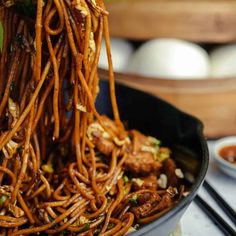 noodles being lifted from a pan by chopsticks with sauce and eggs in the background