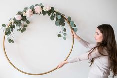 a woman is holding up a gold hoop with flowers on it and greenery in the middle