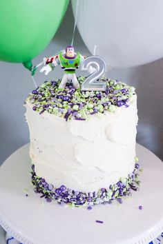 a white cake with purple and green sprinkles is topped with a toy figure