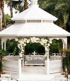 a white gazebo with flowers and greenery on the sides is surrounded by palm trees