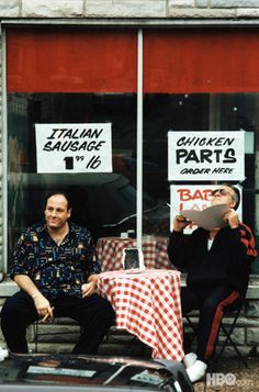 two men sitting on chairs in front of a restaurant with signs reading thai and english parts