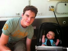 a man kneeling down next to a baby in a stroller and smiling at the camera