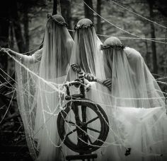 three ghostlyly dressed women standing next to a wheel in the woods with long veils over their heads