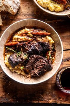 a bowl filled with meat and potatoes on top of a wooden table next to bread