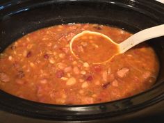 a wooden spoon in a black crock pot filled with beans and meats, ready to be cooked