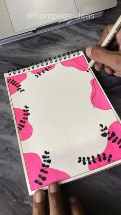 a person is drawing on a notebook with pink and black flowers, while another hand holds a pencil