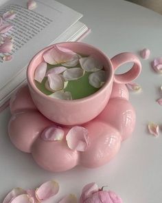 a pink cup filled with water surrounded by petals on top of a table next to an open book