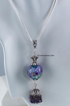 "I have created this one-of-a-kind 'Eastern Iris' pendant necklace from lampwork and dichroic round focal bead containing glass flowers, silver plated brass and bead tassel, silver plated pewter lotus bead, silver plated brass and pewter bead caps, bale and jewellery wire, silver plated brass double snake chain with lobster clasp Alt Text Necklace of lampwork and dichroic round focal bead of glass flowers, silver plated brass and bead tassel, silver plated pewter lotus bead, silver plated brass Silver Glass Flower Pendant Jewelry, Silver Czech Glass Round Necklace, Silver Murano Glass Necklace For Jewelry Making, Artistic Silver Jewelry With Round Beads, Round Silver Czech Glass Necklace, Silver Murano Glass Round Pendant Necklace, Silver Murano Glass Round Pendant Jewelry, Silver Murano Glass Round Necklace, Iris Pendant