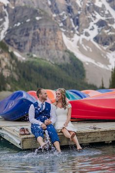 a man and woman sitting on a dock next to canoes in the water with snow capped mountains behind them