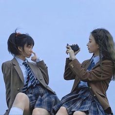 two young women dressed in school uniforms sitting on top of a rock, one holding a camera