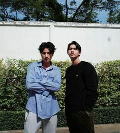 two young men standing next to each other in front of a white wall and bushes