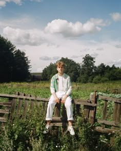 a young man sitting on top of a wooden fence in a field with tall grass