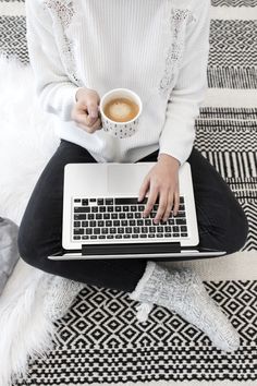 a woman is sitting on the floor with her laptop and holding a cup of coffee