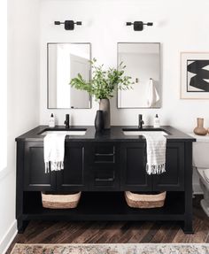 a black vanity with two sinks and mirrors in a white walled bathroom that has a rug on the floor next to it