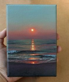 a person holding up a small painting of the sun setting over the ocean with waves