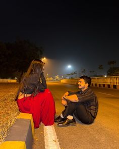 a man and woman sitting on the side of a road next to each other at night