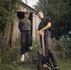 two young men standing next to each other holding guitars in front of a wooden shed