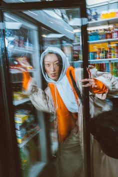 a woman standing in front of a refrigerator filled with food and drinks, wearing an orange hoodie