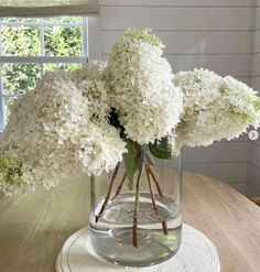 white hydrangeas in a glass vase on a table with a window behind them