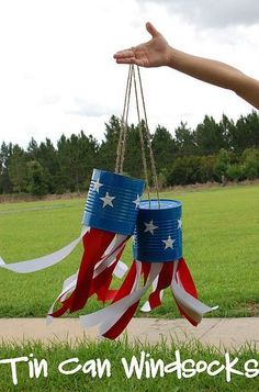 a hand is holding two patriotic tin can wind chimes in the grass with trees in the background