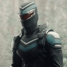 a man dressed in black and blue armor standing in front of trees with his hands on his hips