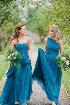 two bridesmaids in blue dresses walking down a path with greenery on either side