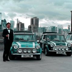 a man in a tuxedo standing next to two old cars with skyline in the background