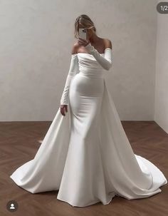 a woman in a white dress taking a selfie with her cell phone while wearing a long sleeved wedding gown