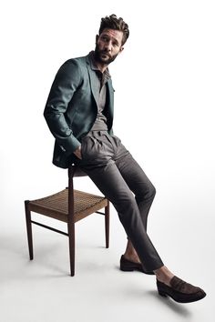 a man sitting on top of a wooden chair next to a white wall with his legs crossed