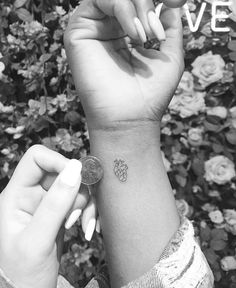 a person with a small pineapple tattoo on their wrist holding a coin in front of some flowers