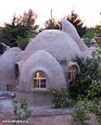 an unusual looking house in the middle of some trees and bushes, with lights on