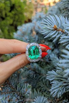 a woman's hand holding an emerald and diamond ring on top of a pine tree