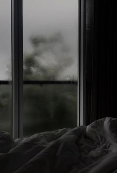 a bed sitting in front of a window next to a lush green forest under a cloudy sky