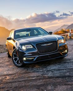 the front end of a black car parked on top of a dirt field with mountains in the background