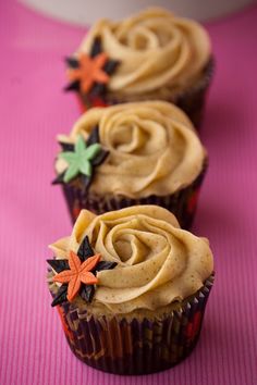 three cupcakes with frosting sitting on a pink tablecloth and one has an orange flower