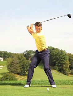 a man swinging a golf club on top of a lush green field with trees in the background