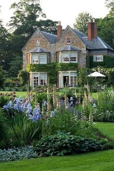 a large house surrounded by lush green grass and flowers in the foreground, with an abundance of trees and shrubs surrounding it