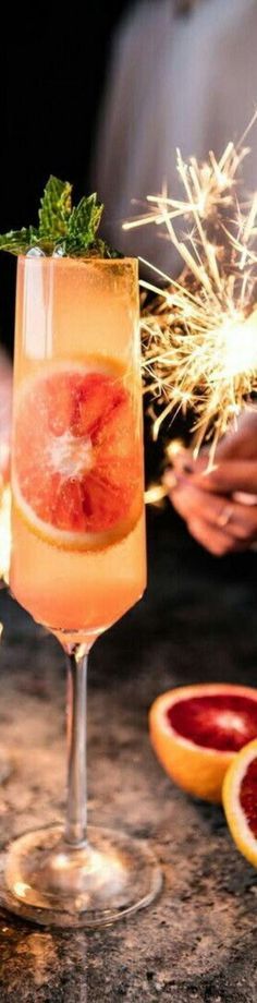 a person holding a sparkler near a glass filled with liquid and garnished with grapefruit