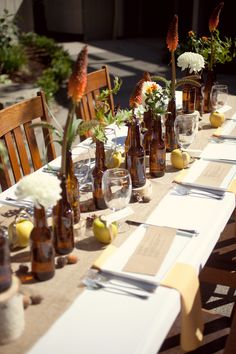 a long table with several bottles and flowers on it, along with place settings for two people