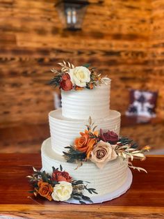 a three tiered white wedding cake with flowers on the top and sides, sitting on a wooden table