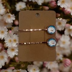 two pairs of blue and white flower hair pins in a cardboard box with flowers behind them