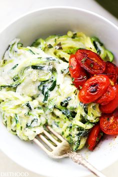 a white bowl filled with pasta and tomatoes on top of a wooden table next to a fork