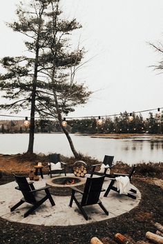 an outdoor fire pit with chairs around it and lights hanging from the trees over the water